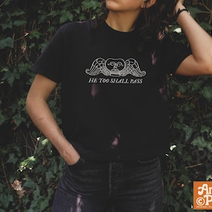 May include: A black short-sleeve t-shirt featuring a white graphic of a winged skull with the text HE TOO SHALL PASS printed underneath. The design is centered on the chest of the garment, which is paired with dark denim jeans.