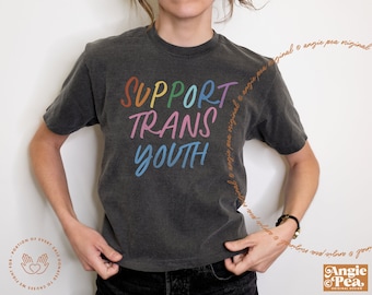 May include: A dark grey t-shirt featuring the text "SUPPORT TRANS YOUTH" in rainbow colours. The shirt has short sleeves and a faded appearance. The design includes the text "angie pea original" and a logo.
