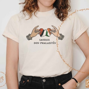 Op de afbeelding: Een wit T-shirt met een afbeelding van twee handen die een hart vasthouden met de Palestijnse en Ierse vlaggen. De tekst "SAOIRSE DON PHALAISTÍN" is onder het hart gedrukt.