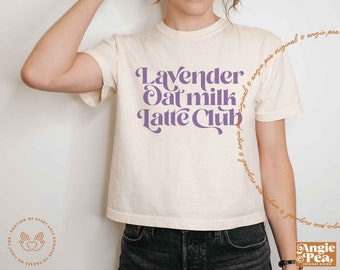 May include: A cream-coloured t-shirt featuring the purple text "Lavender Oat Milk Latte Club" in a retro font. The short-sleeved tee has a crew neck. The design includes the brand name "Angie Pea" in the bottom right corner.