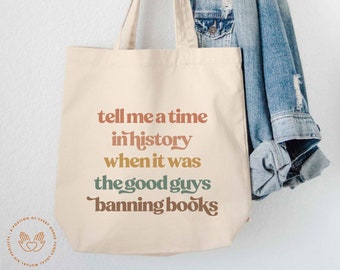 May include: A cream-colored canvas tote bag featuring the text "tell me a time in history when it was the good guys banning books" printed in stacked lines of terracotta, mustard yellow, and dark green, with two fabric handles at the top.