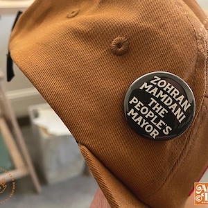 May include: A brown baseball cap with a black and white button pinned to it. The button reads "Zohran Mamdani The People's Mayor" in white text on a black background. The cap is made of a textured fabric.