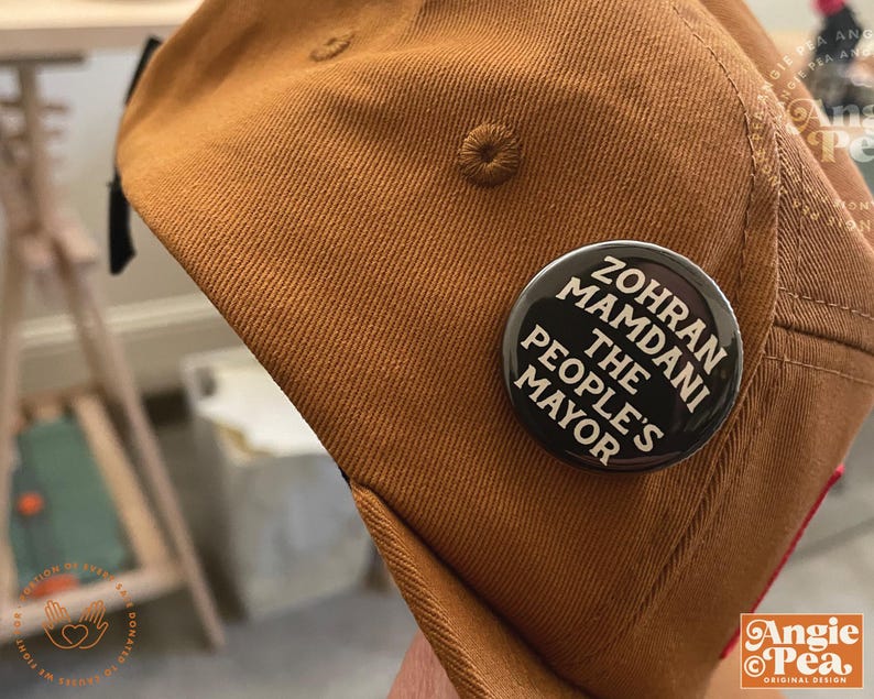 May include: A brown baseball cap with a black and white button pinned to it. The button reads "Zohran Mamdani The People's Mayor" in white text on a black background. The cap is made of a textured fabric.