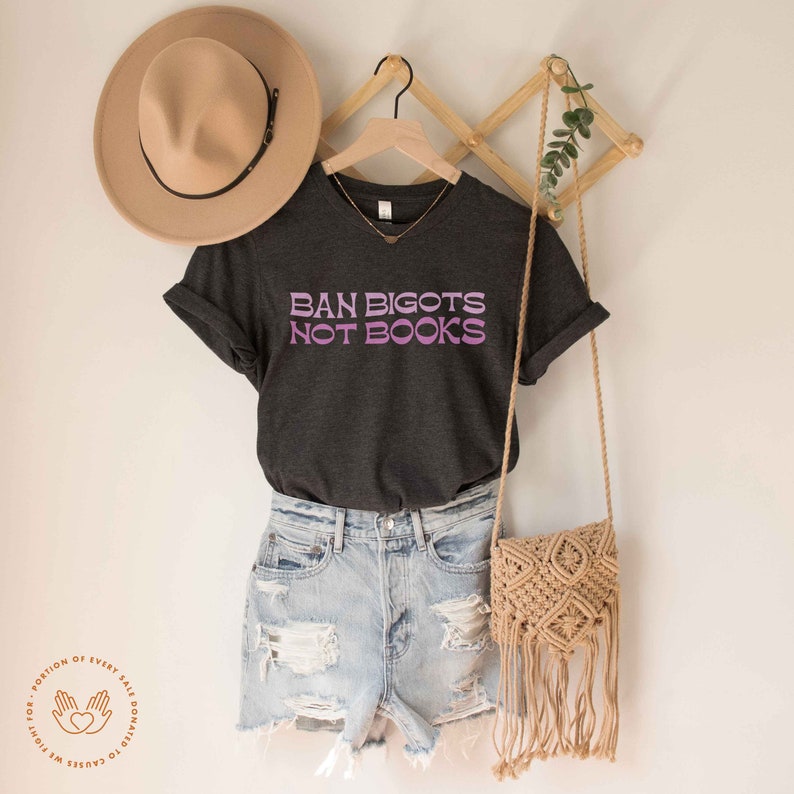 K&ouml;nnte beinhalten: Ein dunkelgraues T-Shirt mit einem lila und wei&szlig;en Aufdruck, der "Ban Bigots Not Books" lautet. Das Shirt h&auml;ngt an einem Holzb&uuml;gel und wird mit Jeans-Shorts und einer Strohtasche gestylt.