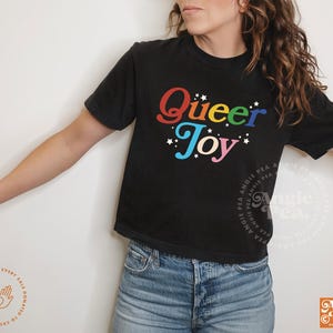 May include: Black t-shirt with the words "Queer Joy" in rainbow colors and white stars. The shirt is paired with blue denim jeans. The design includes the Angie Pea logo.