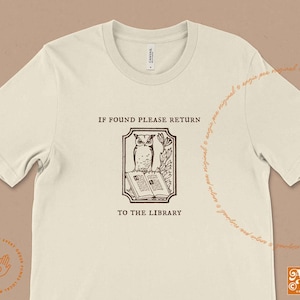 May include: A beige t-shirt with a brown graphic of an owl perched on a book. The graphic is surrounded by a frame and the text "IF FOUND PLEASE RETURN TO THE LIBRARY".