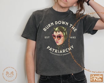 May include: A black cropped t-shirt with a graphic print of a cartoon woman wearing a green devil costume and sunglasses. The text "Burn Down The Patriarchy" is printed on the shirt, along with the text "Est 1692" and "Torch Systems of Oppression".