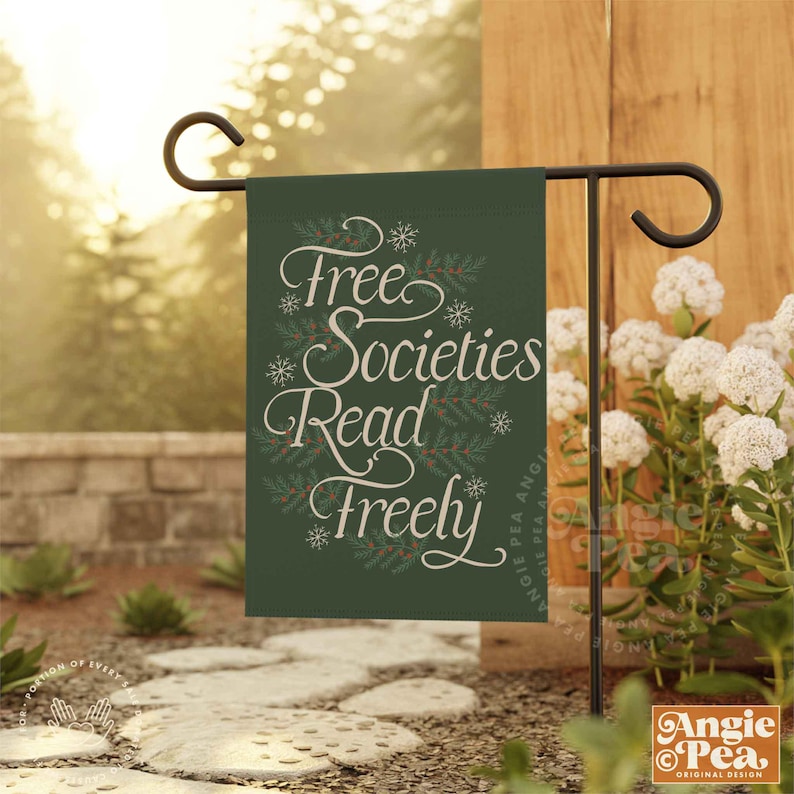 May include: A green garden flag with white text that reads "Free Societies Read Freely". The flag is hanging on a black metal flagpole.