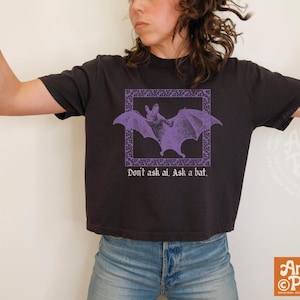 May include: A black short-sleeved t-shirt featuring a purple graphic of a bat inside a decorative square frame. Below the bat, the text reads, "Don't ask ai. Ask a bat." The shirt has a crew neckline and a boxy, cropped fit, paired with blue jeans.