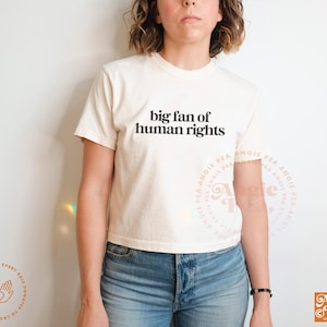 May include: Cream-colored t-shirt with the text "big fan of human rights" in black. The shirt is paired with blue jeans. The image is well-lit, showcasing the shirt's design and the overall casual style.