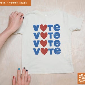 Puede incluir: Una camiseta de manga corta color crema con un estampado gráfico de la palabra VOTE repetida cuatro veces en una fuente azul desgastada. Cada letra O está reemplazada por un corazón rojo. El diseño está centrado en la parte delantera.