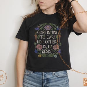 May include: Black cropped t-shirt with a floral design and the text "Continuing to care for others is to resist".