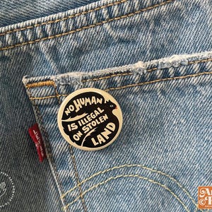 May include: A round, black and white button with the text "NO HUMAN IS ILLEGAL ON STOLEN LAND" is pinned to the back pocket of a pair of blue denim jeans. The jeans have visible stitching and a red Levi's tag.