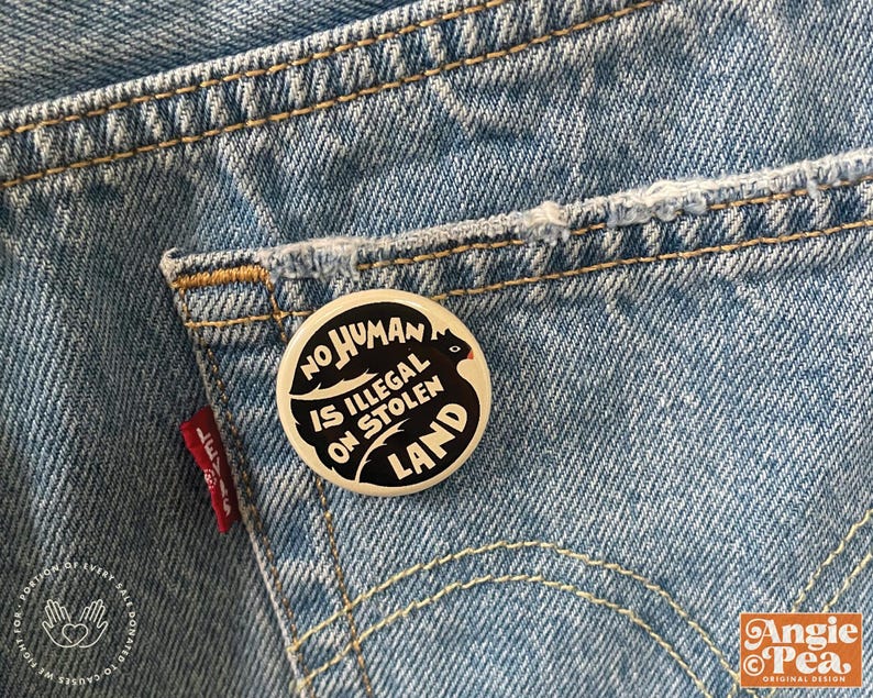 May include: A round, black and white button with the text "NO HUMAN IS ILLEGAL ON STOLEN LAND" is pinned to the back pocket of a pair of blue denim jeans. The jeans have visible stitching and a red Levi's tag.