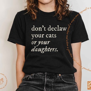 May include: Black t-shirt with white text that reads "don't declaw your cats or your daughters."