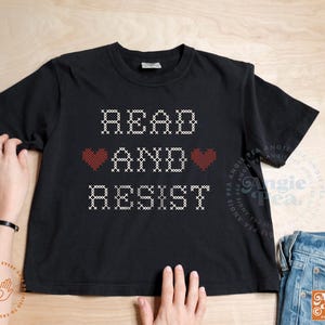 May include: Black t-shirt with the words "READ AND RESIST" in a white, cross-stitch style font. Two red hearts are between the words. The shirt is displayed on a wooden surface with a pair of blue jeans.