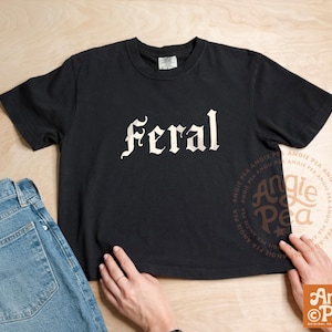 Puede incluir: Una camiseta negra de manga corta con la palabra feral impresa en una fuente gótica blanca en el pecho. La prenda se muestra junto a unos vaqueros azules, exhibiendo un diseño sencillo y casual para el uso diario.