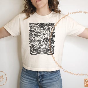 K&ouml;nnte beinhalten: Ein wei&szlig;es T-Shirt mit einem schwarzen floralen Design und dem Text "Fragile Like A Bomb".