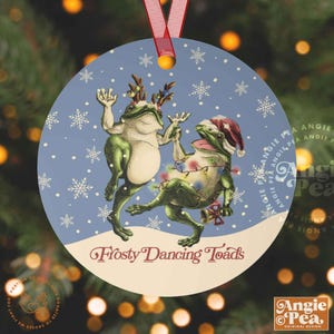 May include: Round Christmas ornament featuring two green frogs dancing in a winter scene. One frog wears a Santa hat and is wrapped in Christmas lights. The other frog has antlers. The ornament has the text "Frosty Dancing Toads" and is suspended by a red ribbon.