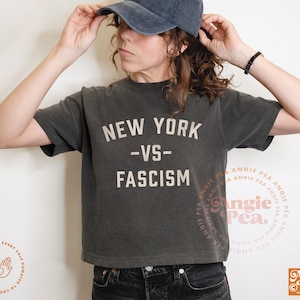 Puede incluir: Camiseta gris oscuro con el texto blanco "NEW YORK -VS- FASCISM". La camiseta es de estilo corto y se combina con una gorra de béisbol de mezclilla y vaqueros negros. La camiseta tiene un aspecto vintage y desgastado.