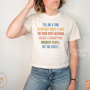 May include: Cream-colored t-shirt with the text "TELL ME A TIME IN HISTORY WHEN IT WAS THE GOOD GUYS WEARING MASKS & KIDNAPPING INNOCENT PEOPLE OFF THE STREET." in various colors. The shirt is paired with blue jeans.