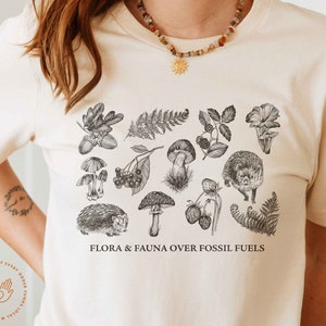 May include: A white t-shirt with a black and white illustration of various flora and fauna, including mushrooms, berries, ferns, and hedgehogs. The text "FLORA & FAUNA OVER FOSSIL FUELS" is printed on the shirt.
