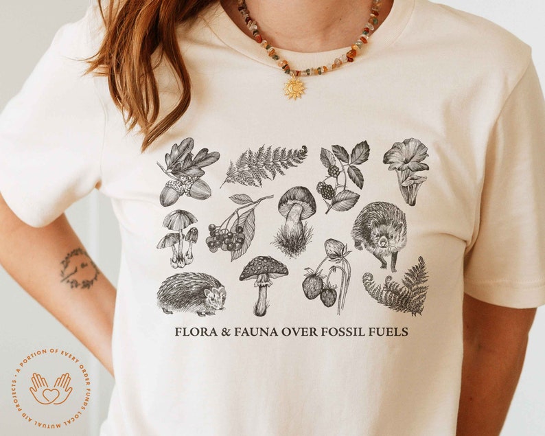 May include: A white t-shirt with a black and white illustration of various flora and fauna, including mushrooms, berries, ferns, and hedgehogs. The text "FLORA & FAUNA OVER FOSSIL FUELS" is printed on the shirt.