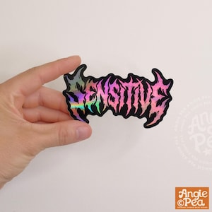 May include: A die-cut sticker featuring the word SENSITIVE written in a jagged, heavy metal-style font. The lettering displays a vibrant holographic finish that shifts between shades of pink, purple, blue, and green against a black outline.