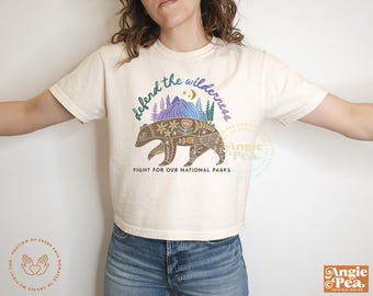 May include: A white cropped t-shirt with a brown bear illustration and the text "defend the wilderness" in a colorful font. The text "FIGHT FOR OUR NATIONAL PARKS" is below the bear.