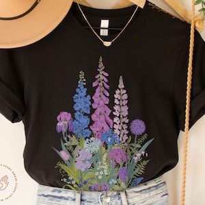 Puede incluir: Camiseta negra con estampado floral con flores moradas, azules y verdes. El texto "A portion of every order funds local mutual aid projects." est&aacute; impreso en la esquina inferior izquierda de la camiseta.