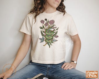 May include: A white t-shirt with a green snake and flowers printed on the front. The snake is wrapped around a banner that says "Protect Trans Folks".
