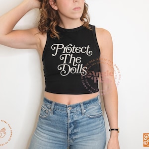 May include: Black crop top with the text "Protect The Dolls" in white script. The top is paired with blue denim jeans. The image also includes the Angie Pea logo and a circular graphic with text.