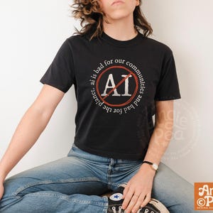 May include: Black t-shirt with the text "ai is bad for our communities and bad for the planet." The text is in a white circular design with a red line through the letters "AI". The shirt is worn with blue jeans.