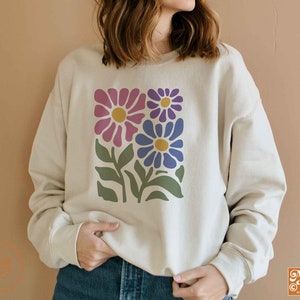 May include: A cream-colored sweatshirt with a floral design featuring pink, purple, and green flowers. The flowers are stylized and have a retro feel.