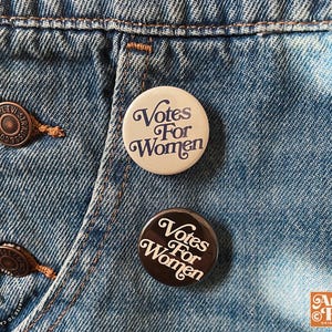 May include: Two round buttons with the text "Votes For Women" in a vintage font. One button is cream-colored with navy text, the other is dark brown with cream text. The buttons are pinned to a blue denim garment.