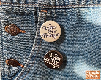 Puede incluir: Dos botones redondos con el texto "Votes For Women" en una fuente vintage. Un botón es de color crema con texto azul marino, el otro es marrón oscuro con texto crema. Los botones están prendidos a una prenda de mezclilla azul.