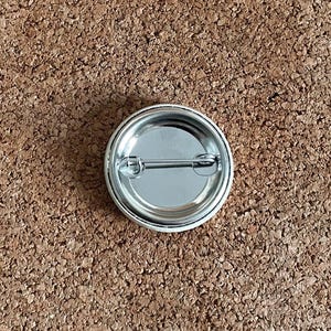May include: A round, silver-colored pinback button with a safety pin on the back. The button has a shiny, metallic finish and a simple, classic design. The button is shown against a brown, textured background.
