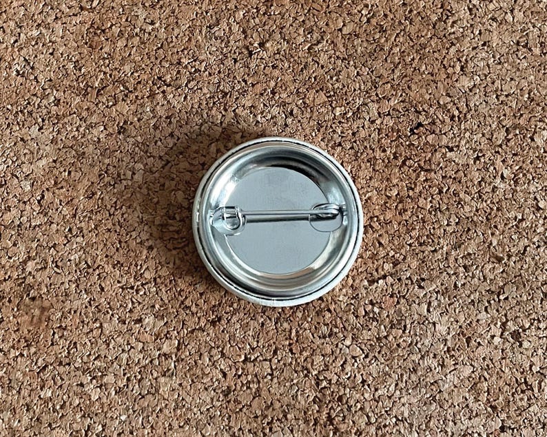 May include: A round, silver-colored pinback button with a safety pin on the back. The button has a shiny, metallic finish and a simple, classic design. The button is shown against a brown, textured background.