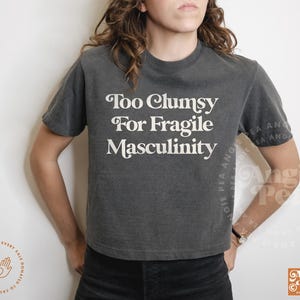May include: A dark gray t-shirt with the text "Too Clumsy For Fragile Masculinity" in white. The shirt has short sleeves and a crew neck. The shirt is worn by a person.