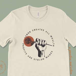 May include: A beige t-shirt with a black and red graphic design. The graphic features a fist holding a rose with the text "Labor Creates All Wealth Profits Are Stolen Wages".