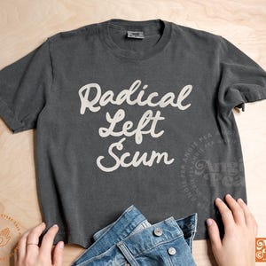 May include: Dark gray t-shirt with the words "Radical Left Scum" in white script. The shirt is paired with blue denim jeans. The image is on a light wood surface.