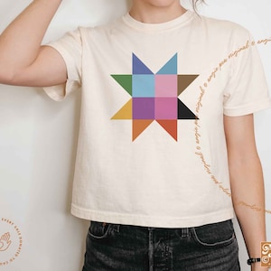 May include: A white cropped t-shirt with a colorful patchwork star design. The text "original o angie pea niginal o angie pea niginal o angie pea niginal" is written in a cursive font around the star. The text "Angie Pea, Original Design" is in the bottom right corner of the image.