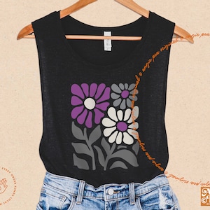 May include: Black sleeveless shirt featuring a floral design in purple, gray, and white. The graphic includes stylized flowers and leaves. The shirt is displayed on a wooden hanger against a neutral background. The text "Angie Pea Original Design" is visible.