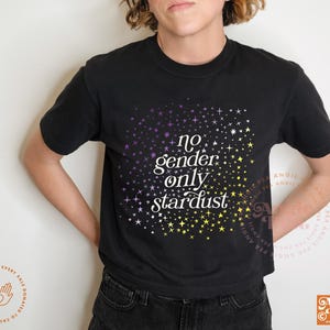 May include: Black t-shirt with the text "no gender only stardust" in white, with a starry design in purple and yellow. The shirt has short sleeves and a crew neck.