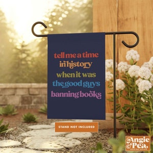 May include: A blue garden flag with the text "tell me a time in history when it was the good guys banning books" in rainbow colors. The text is on a dark blue background. The flag is hanging from a black metal stand. The stand is not included.