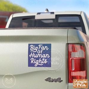 May include: A square blue and white bumper sticker with the text "Big Fan of Human Rights" is affixed to the back of a light green truck. The sticker has a gradient background and a white script font. The Angie Pea logo is in the bottom right corner.