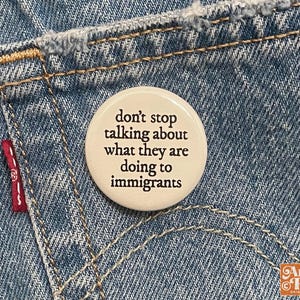 May include: A round, off-white button with the text "don't stop talking about what they are doing to immigrants" in black. The button is pinned to a blue denim pocket with a red Levi's tag.