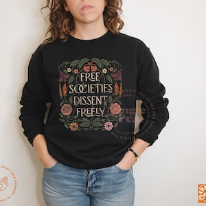 May include: Black sweatshirt with the text "FREE SOCIETIES DISSENT FREELY" in an ornate floral design. The design includes flowers in shades of pink, orange, and yellow. The sweatshirt has long sleeves.