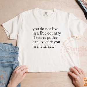 May include: Cream-colored t-shirt with the text "you do not live in a free country if secret police can execute you in the street." The shirt is paired with blue jeans. The image is taken from a top-down perspective.