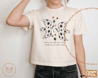 Könnte beinhalten: Ein weißes, bauchfreies T-Shirt mit einer schwarzen und grünen Grafik von zwei stilisierten Tauben, die sich gegenüberstehen. Die Tauben sind von kleinen Sternen umgeben. Der Text "Until we are all free, none of us are free." ist unter den Tauben gedruckt.